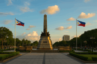 A wide sunset view of the Rizal Monument at Rizal Park, framed by two Philippine flags and surrounded by trees, pathways, visitors, and a warm golden sky.